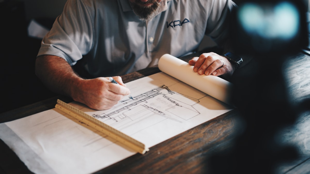 A person in a grey shirt sketches architectural plans on a large paper with a pencil, using a ruler, at a wooden desk.